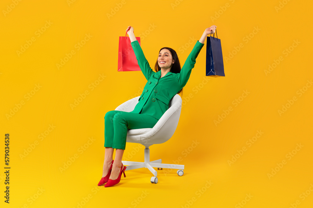 Fototapeta premium Full body length shot of contented shopaholic woman sitting on chair and raising hands with shopping bags, free space