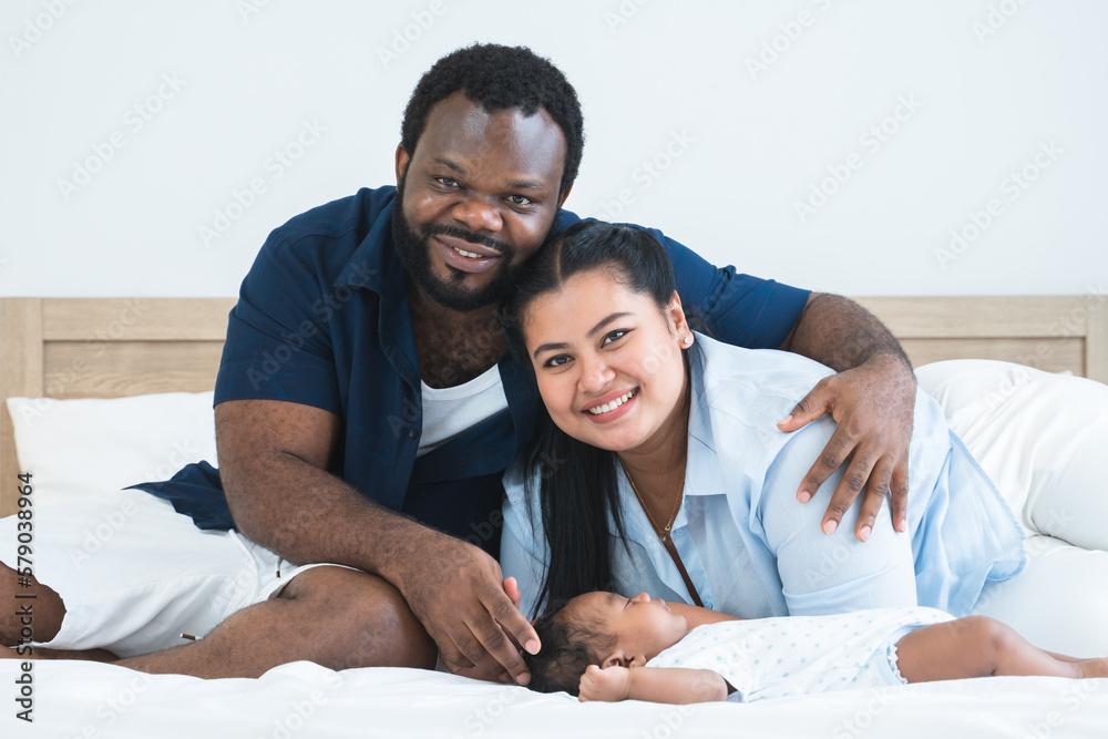 Fototapeta premium African father and Asian mother touching and lull cute newborn baby sleeping lying on bed at home, parent smiling looking at camera. Multiracial family bonding and child care concept