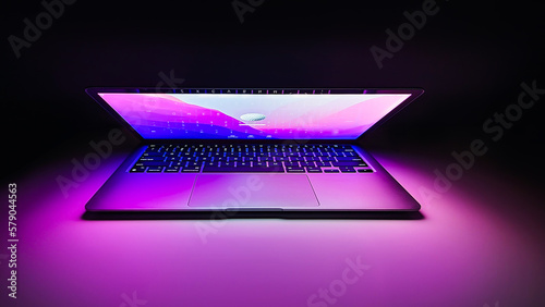 Laptop with half lid open on a table lit with colorful purple and blue desktop screen wallpaper in a dark room	
 photo