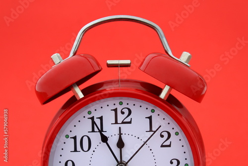 A red alarm clock stands on a red background. The time shows five minutes to twelve.