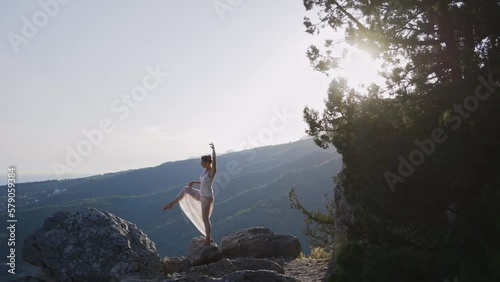 Young woman in long white clothes standing in yoga poses, balancing on one leg on rocky cliff edge under bright summer morning sunlight, Tuladandasana, stick yoga pose, unity with nature concept photo