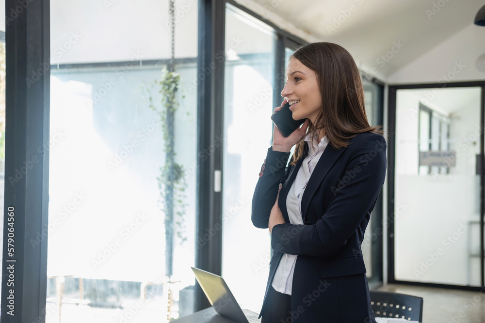 A stylish white businesswoman in a black suit standing inside the office in hands with a mobile phone, tablet, and laptop waiting for customers to contact, business people concept. A stylish white businesswoman in a black suit standing inside the office in hands with a mobile phone, tablet, and laptop waiting for customers to contact, business people concept.