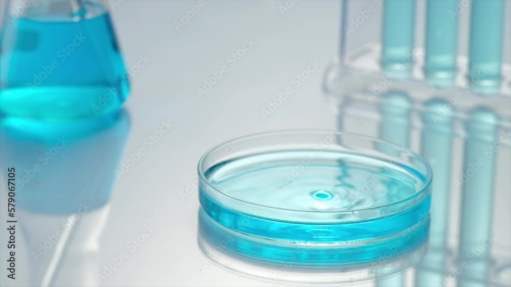 Slow motion of transparent blue fluid dripping into the glass petri dish. Chemical laboratory concept with erlenmeyer flask, tests tube and petri dish on light background. Slow motion of transparent blue fluid dripping into the glass petri dish. Chemical laboratory concept with erlenmeyer flask, tests tube and petri dish on light background.