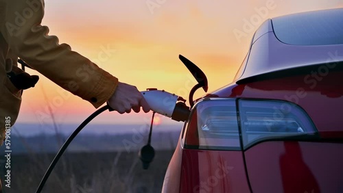 Close view of a man plugging charging cable into a parked red electric car at sunset photo