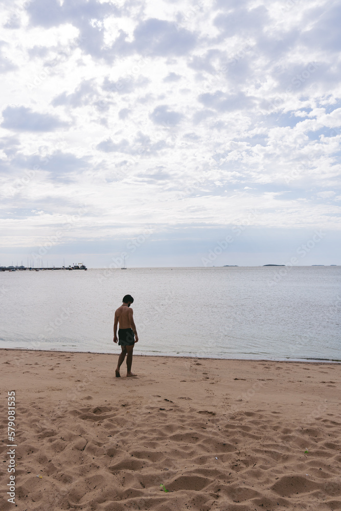 Vertical photo of a young latin man walking towards the water on a sandy beach on a cloudy day. Copy space.