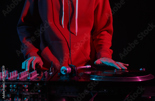 Cool young DJ in red hoodie scratching vinyl records on turntables in night club. Hands of a hip hop disc jockey playing music on stage