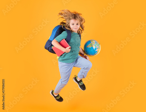 Full length of excited kid jumping. Back to school. School child in school uniform with bagpack and globe jump. School children jumping on studio isolated yellow background.
