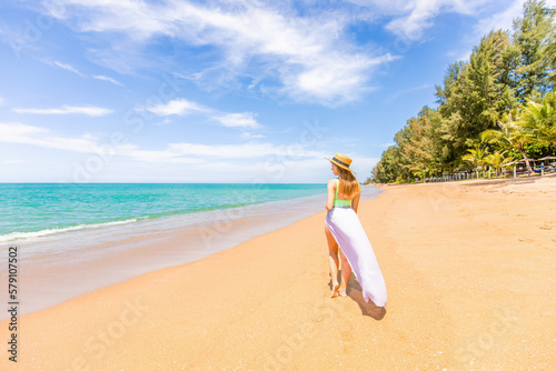 Traveller woman in swimwear enjoys tropical beach vacation. Summer lifestyle of pretty happy young girl walking at ocean coastline. Tropical island beach with clear water. Vacation at Asia, Thailand