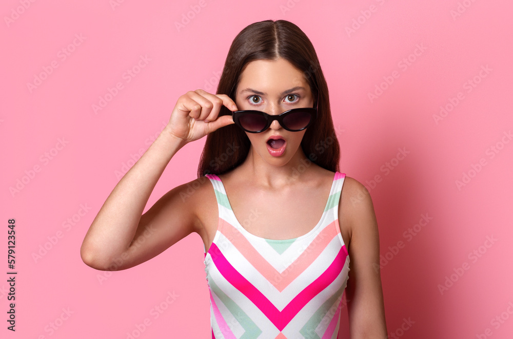amazed surprised teen girl with open mouth taking off sunglasses expressing surprise, dressed in colourful one-piece swimsuit isolated on pink background, summer vacation holidays amazed surprised teen girl with open mouth taking off sunglasses expressing surprise, dressed in colourful one-piece swimsuit isolated on pink background, summer vacation holidays