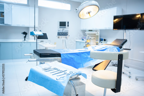 Interior of modern operating room with selective focus of empty doctor s chair beside surgical instruments set on blue cloth  with an empty operating bed with surgical light shining down in background