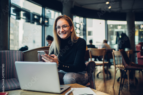 Young woman laughing while using cellphone in cafe and working on netbook