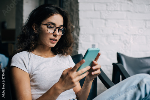 Caucasian female blogger in optical eyewear for vision protection using cellphone technology for social networking in social media, millennial woman with smartphone making online booking and banking