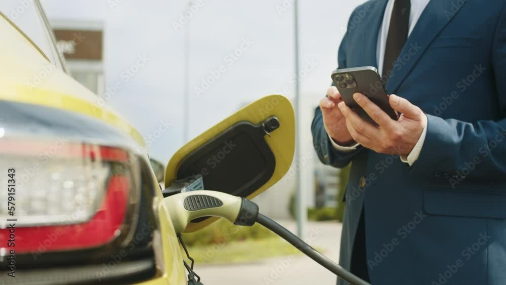 Male unplugging in power cord to electric car using app on smartphone. Businessman charging electric car at outdoor charging station Unrecognizable man unplugging electric car from charging station Male unplugging in power cord to electric car using app on smartphone. Businessman charging electric car at outdoor charging station Unrecognizable man unplugging electric car from charging station