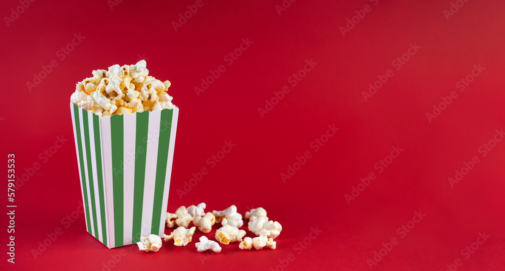Green white striped carton bucket with tasty cheese popcorn, isolated on red background. Box with scattering of popcorn grains. Fast food, movies, cinema and entertainment concept. Green white striped carton bucket with tasty cheese popcorn, isolated on red background. Box with scattering of popcorn grains. Fast food, movies, cinema and entertainment concept.