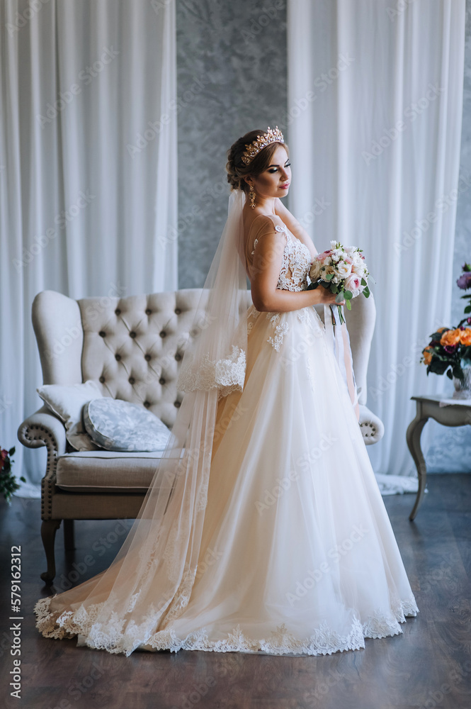 A beautiful bride model blonde in a white long lace dress with a crown, a diadem with a bouquet in her hands stands against the backdrop of a sofa in the studio. Wedding photography, portrait. A beautiful bride model blonde in a white long lace dress with a crown, a diadem with a bouquet in her hands stands against the backdrop of a sofa in the studio. Wedding photography, portrait.