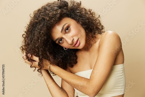 Woman applies cream and balm to her curly hair, the concept of protection and care, a healthy look, a smile with teeth on a beige background