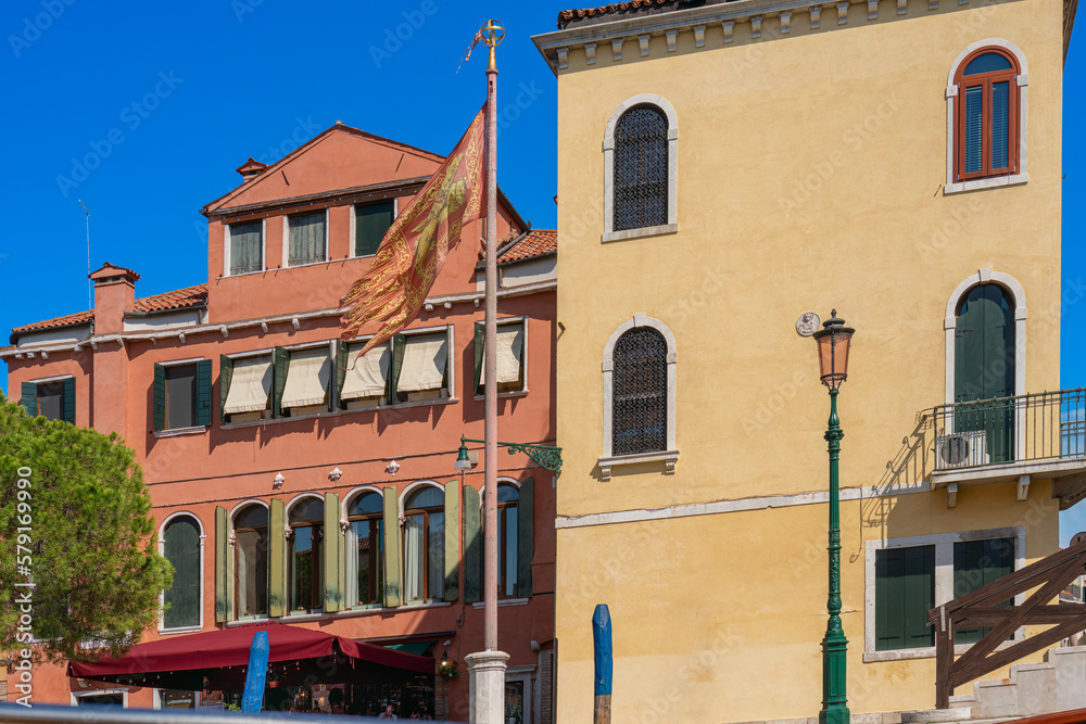Venetian flag on a high pole against the background of the walls of old ancient houses with high windows and wooden shutters, blue sky, summer sunny day, authentic Italian architecture, real world