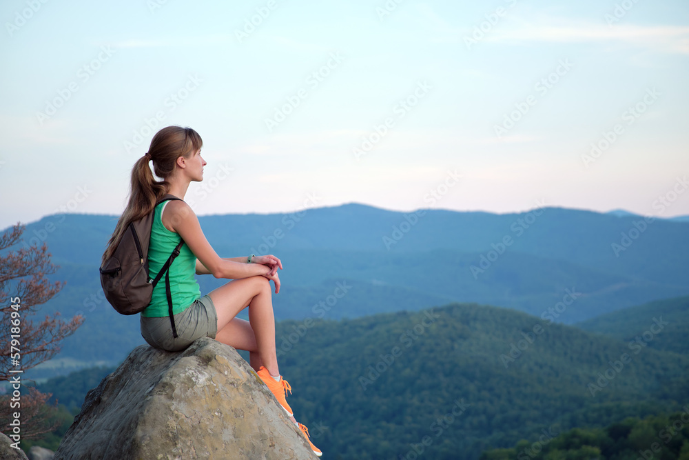 Young woman hiker sitting alone on rocky mountain enjoying view of evening nature on wilderness trail. Active way of life concept