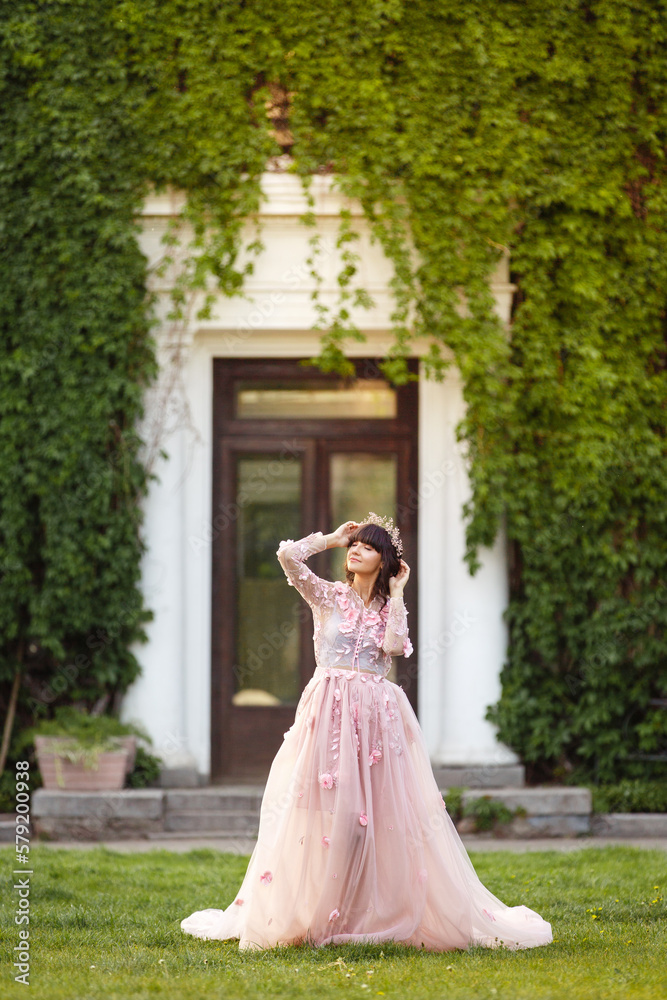 Fototapeta premium Beautiful woman in flying airy pink dress on background of door overgrown with wild grapes. Pretty young caucasian girl plays with her dress. Girl dance in flowing dress