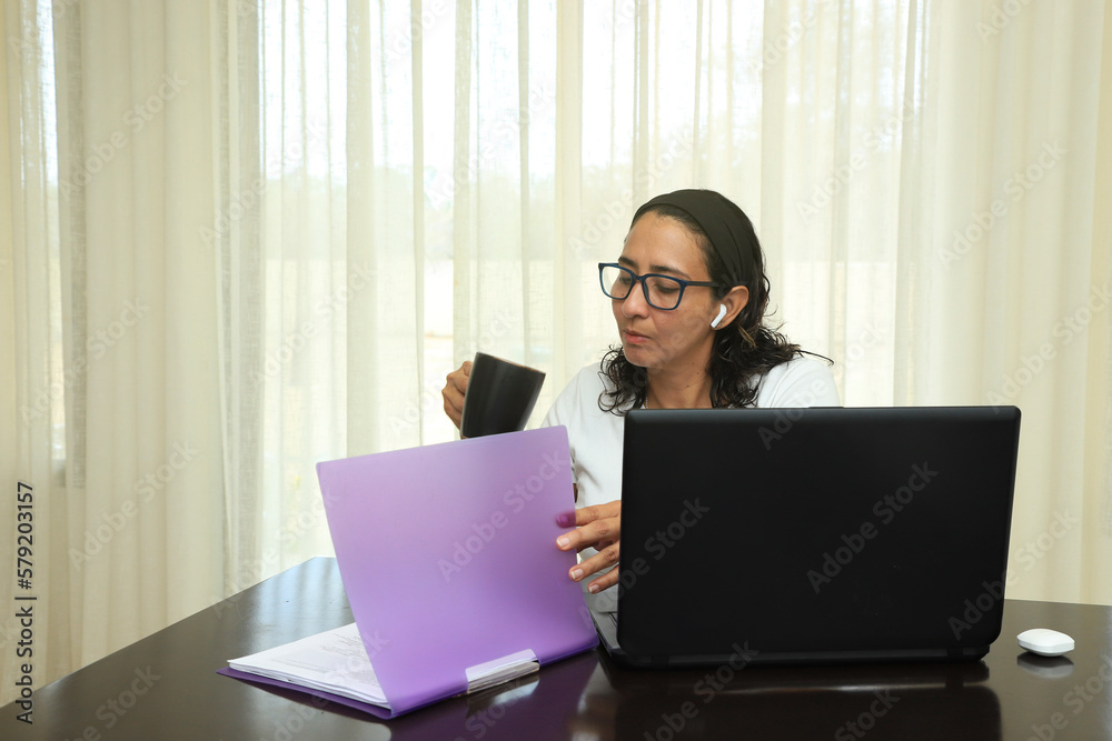 Black-haired Caucasian woman with glasses reading her notes while holding a cup of coffee in front of her computer. Woman from home sitting at table and concentrating in front of computer. Black-haired Caucasian woman with glasses reading her notes while holding a cup of coffee in front of her computer. Woman from home sitting at table and concentrating in front of computer.