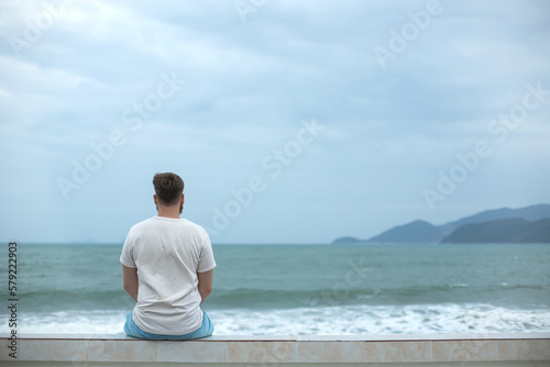 Wallpaper Mural Young calm sad serious man is sitting on embankment near sea, ocean on beach, th