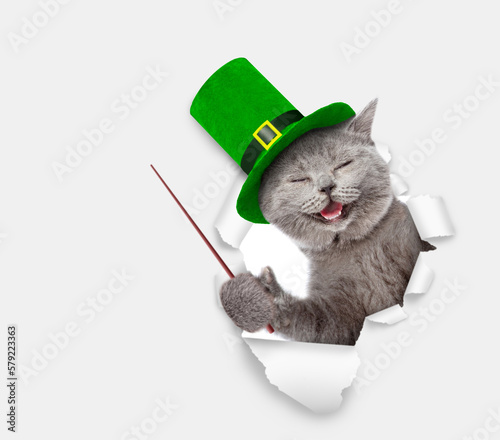 Saint Patricks Day concept. Funny cat wearing hat of the leprechaun looks through the hole in white paper and points away on empty space. isolated on white background. Space for text