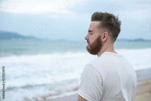 Portrait of young handsome bearded pensive man with beard is enjoy walk on summer beach near sea, breathe deep fresh air in tropical country. Summer vacation.
