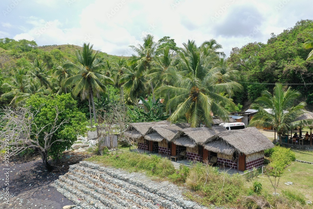 Panorama drone shot of a green space with thatched huts enclosed by rainforest on the site of Pantai Batu Biru, the Blue Stone Beach.