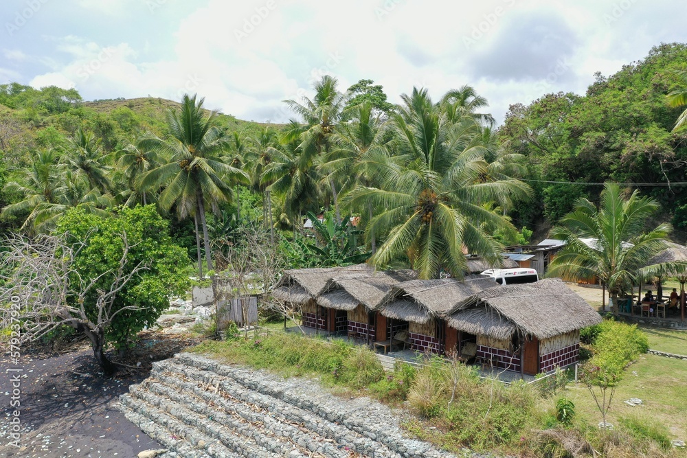 Panorama drone shot of a green space with thatched huts enclosed by rainforest on the site of Pantai Batu Biru, the Blue Stone Beach. Panorama drone shot of a green space with thatched huts enclosed by rainforest on the site of Pantai Batu Biru, the Blue Stone Beach.