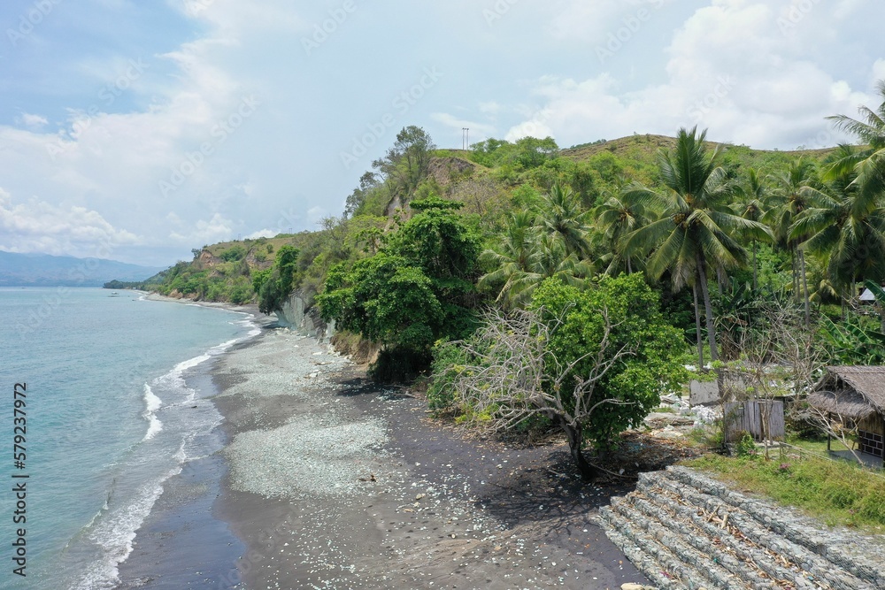 Panorama drone shot sideways along the beach Pantai Batu Biru, the Blue Stone Beach, on the right the stand with trees and hills, on the left the sea.