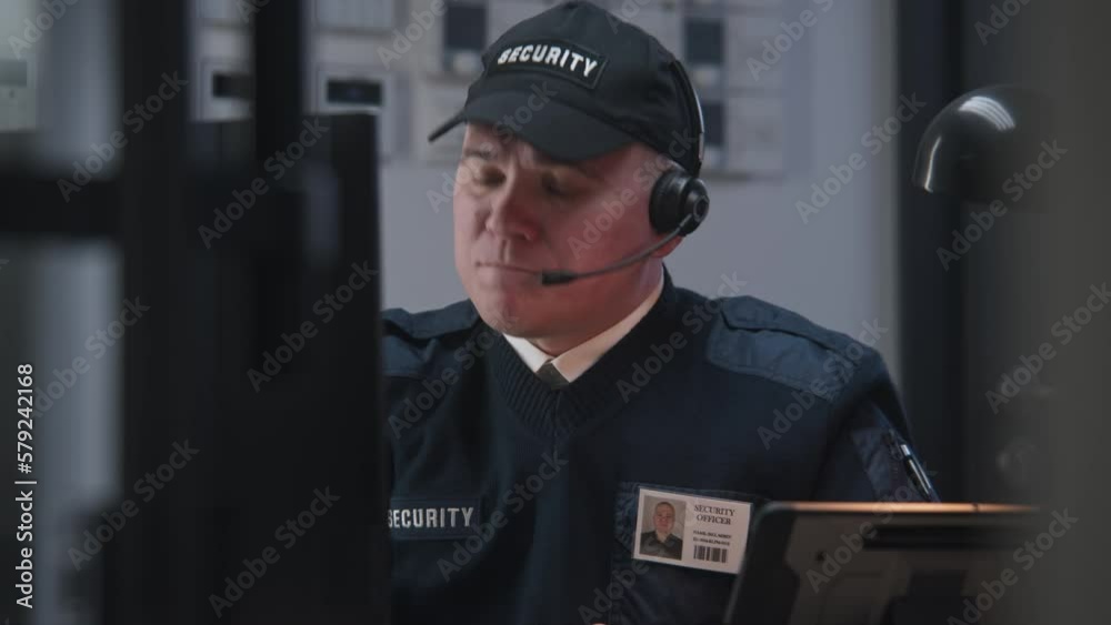 Serious security officer in headphones sits on workplace and looks view from security camera displayed on monitor. Man talks with coworker by walkie talkie. Tracking systems. CCTV technology. Zoom in. Serious security officer in headphones sits on workplace and looks view from security camera displayed on monitor. Man talks with coworker by walkie talkie. Tracking systems. CCTV technology. Zoom in.