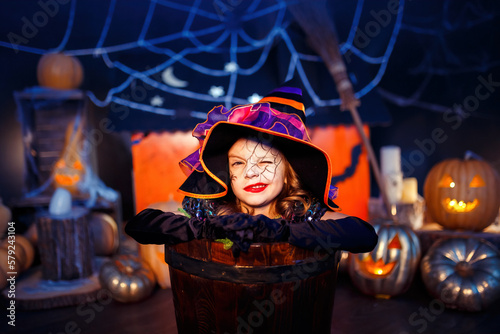 Portrait of winking little girl in witch costume peeks out of stupa on halloween decor background