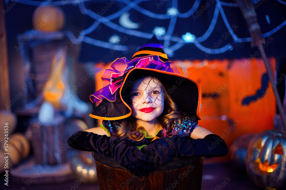 Portrait of little girl in witch costume peeks out of stupa on the background of halloween decor