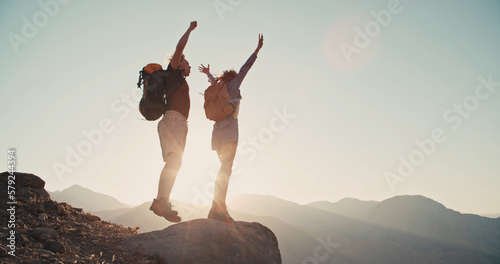 Travelers, man and a woman with backpack jumping on the stone of mountain , raise their hands in air and enjoy victory, beautiful sun and landscape. Free tourists travel. business teamwork
