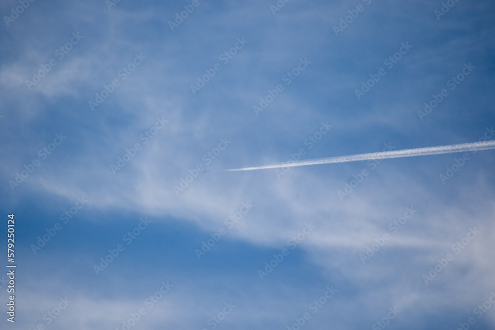 Vapor trail from a jet flying high overhead dissipates into the thin white clouds in a blue sky Vapor trail from a jet flying high overhead dissipates into the thin white clouds in a blue sky