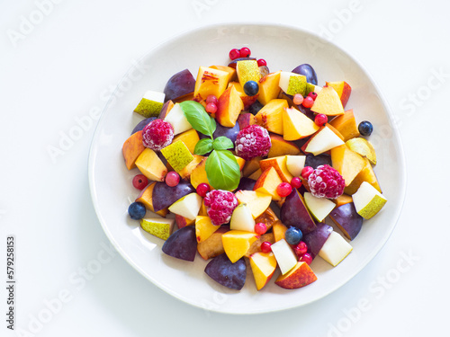 Delicious fruit salad with fresh plum  peach  nectarine  pear and frozen raspberries  decorated with green basil on white background. Top view. Healthy eating. Copy space. Vegan cuisine. Dietary menu.