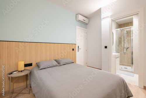 Double bed with gray linen, small bedside table and wooden slats on the wall overlooking compact shower room. Concept of small multifunctional apartment. Copyspace