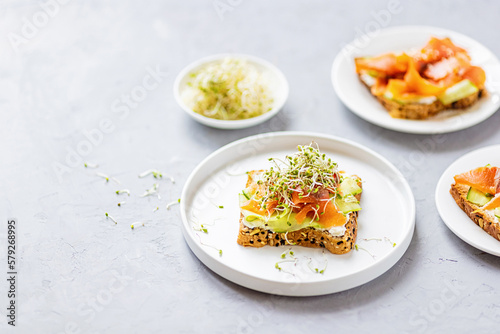 Flat lay top view of healthy sandwich with smoked salmon, cucumber, cream cheese and fresh microgreens alfalfa sprouts on white plate on gray concrete background. Healthy lifestyle. Growing sprouts