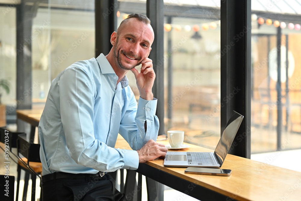 Smiling millennial man using laptop, working remotely in bright coffee shop interior. Business, freelance concept