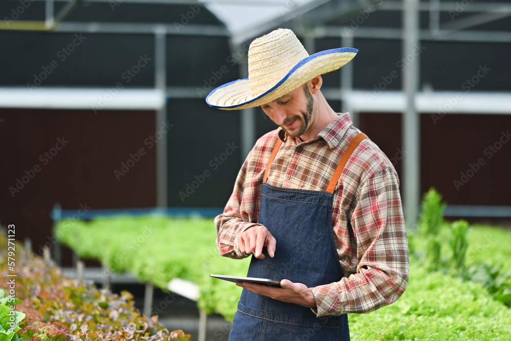 Portrait of farmer standing among his commercial greenhouse and using digital tablet. Agricultural and technology concept