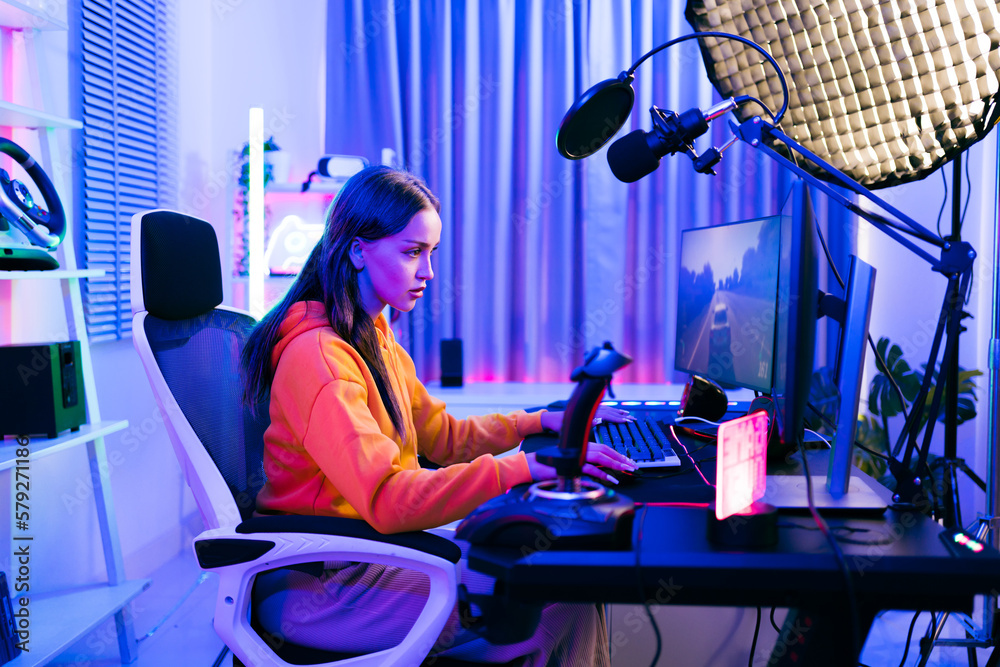 Young Caucasian woman professional gamer wearing yellow hoodie sits on a  chair with a gaming table with pc, keyboard, monitor, microphone. Prepare  for competition, cast gameplay or record a podcast. Stock Photo