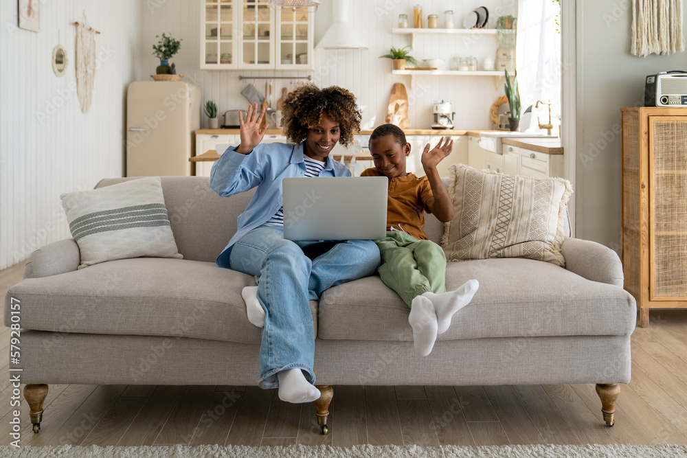 Happy family. African mother and kid son greeting online, waving hands, looking at web camera laptop for video call sitting on couch at home. Smiling mom and child having fun talking in video chat . Happy family. African mother and kid son greeting online, waving hands, looking at web camera laptop for video call sitting on couch at home. Smiling mom and child having fun talking in video chat .