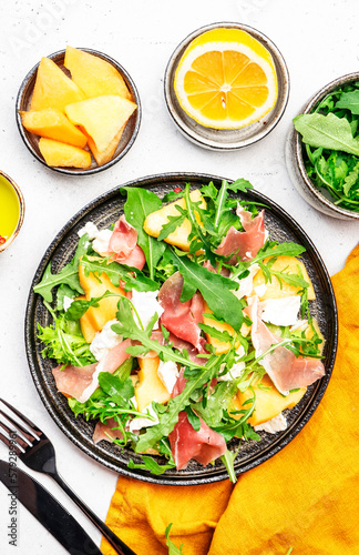 Delicious melon salad with cantaloupe, prosciutto, soft cheese and arugula on white table background, top view