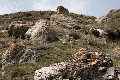 Heap of huge grey boulders with cracks, splits, orange lichen on slope on dim green dry grass meadow, blue sky in highland in summer weather. Mountain valley texture, background, detail.