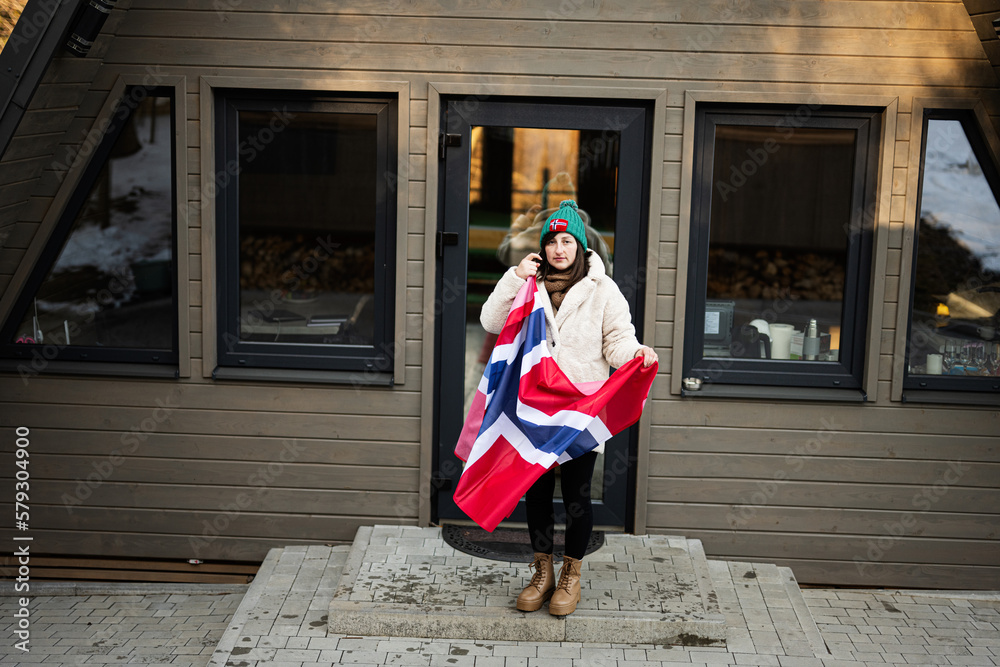 Portrait of woman outside cabin house holding Norway flag. Scandinavian culture, norwegian people.