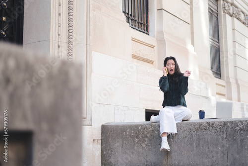 Young Asian girl sitting on the wall at the entrance of the university surprised talking on the phone, next to her is a cup of coffee and a laptop. Lifestyle and technology.