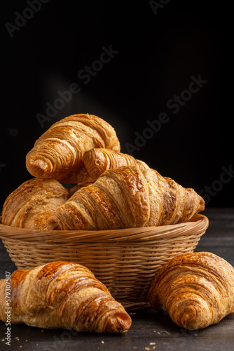 Close-up of croissants in basket on board on dark wooden table with crumbs, selective focus, black background, vertical, with copy space