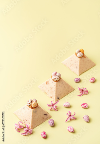 Traditional Easter cottage cheese dessert Paskha decorated with caramel and chocolate candy eggs and spring flowers on pastel yellow background. Modern minimalistic Easter background with copy space.