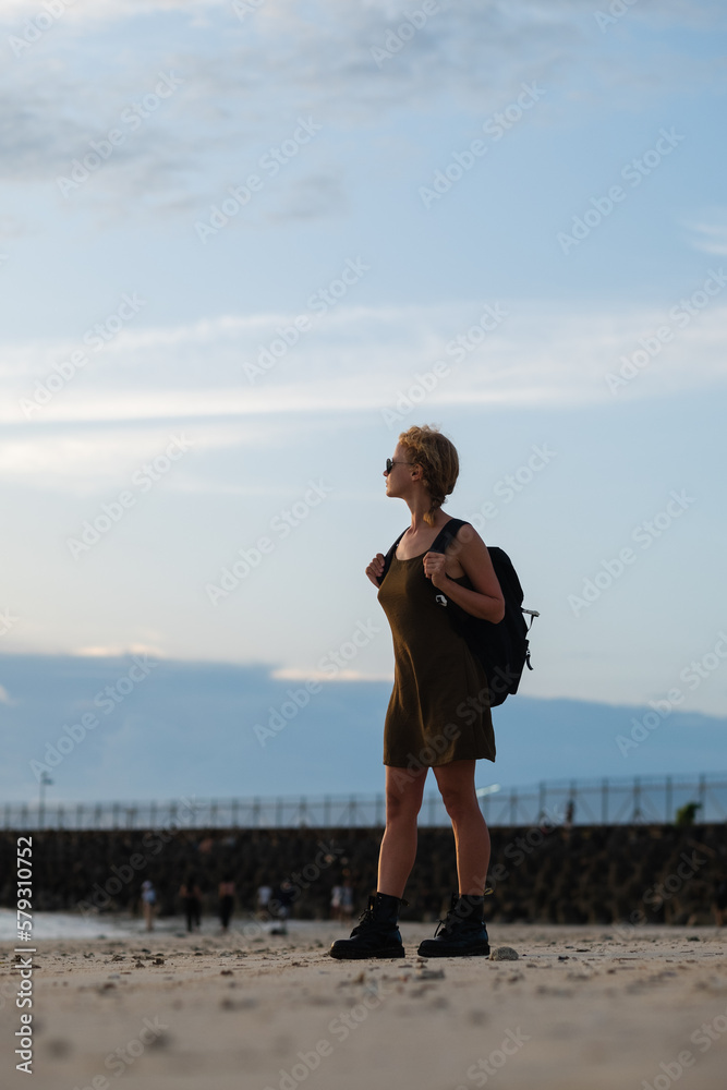 Silhouette of a traveler girl with a backpack, standing by the sea on the beach, meeting the sunset.