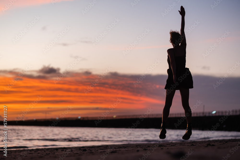A young girl admires the sunset on the ocean, enjoys life, arms outstretched to the sides, rear view.